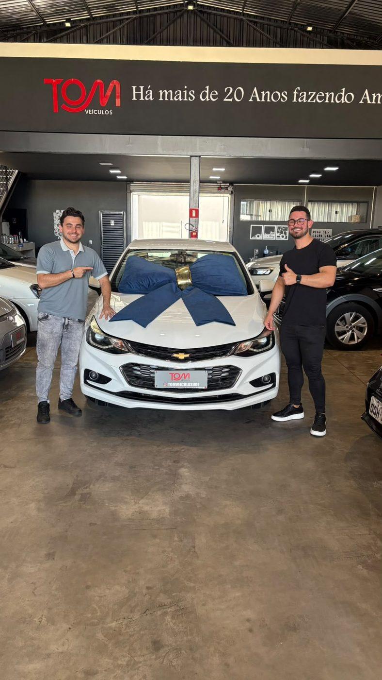 Cliente da Tom Veículos recebendo as chaves do carro novo, com laço de entrega no capô.