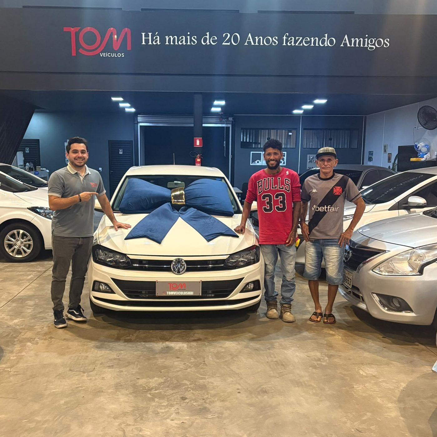 Cliente e equipe da Tom Veículos ao lado do carro entregue com laço decorativo.